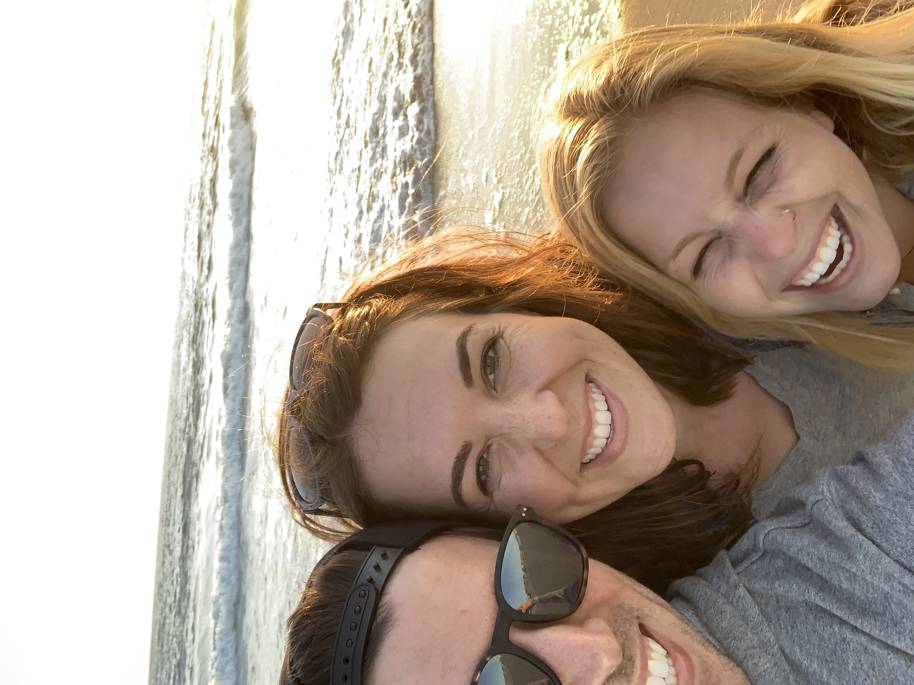 Three people smile together at the beach during golden hour, with ocean
