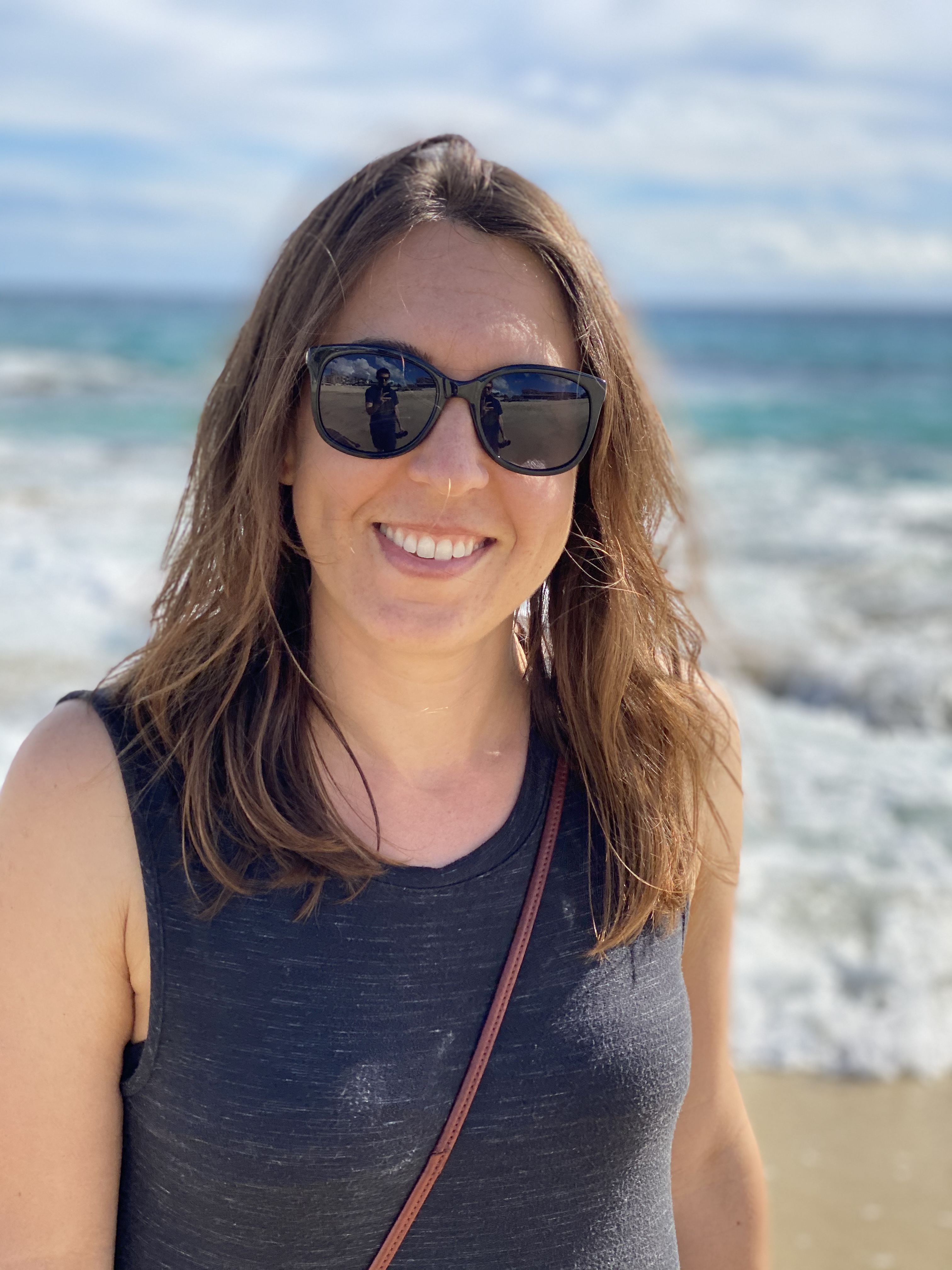 A smiling woman with long brown hair and sunglasses stands on a beach,