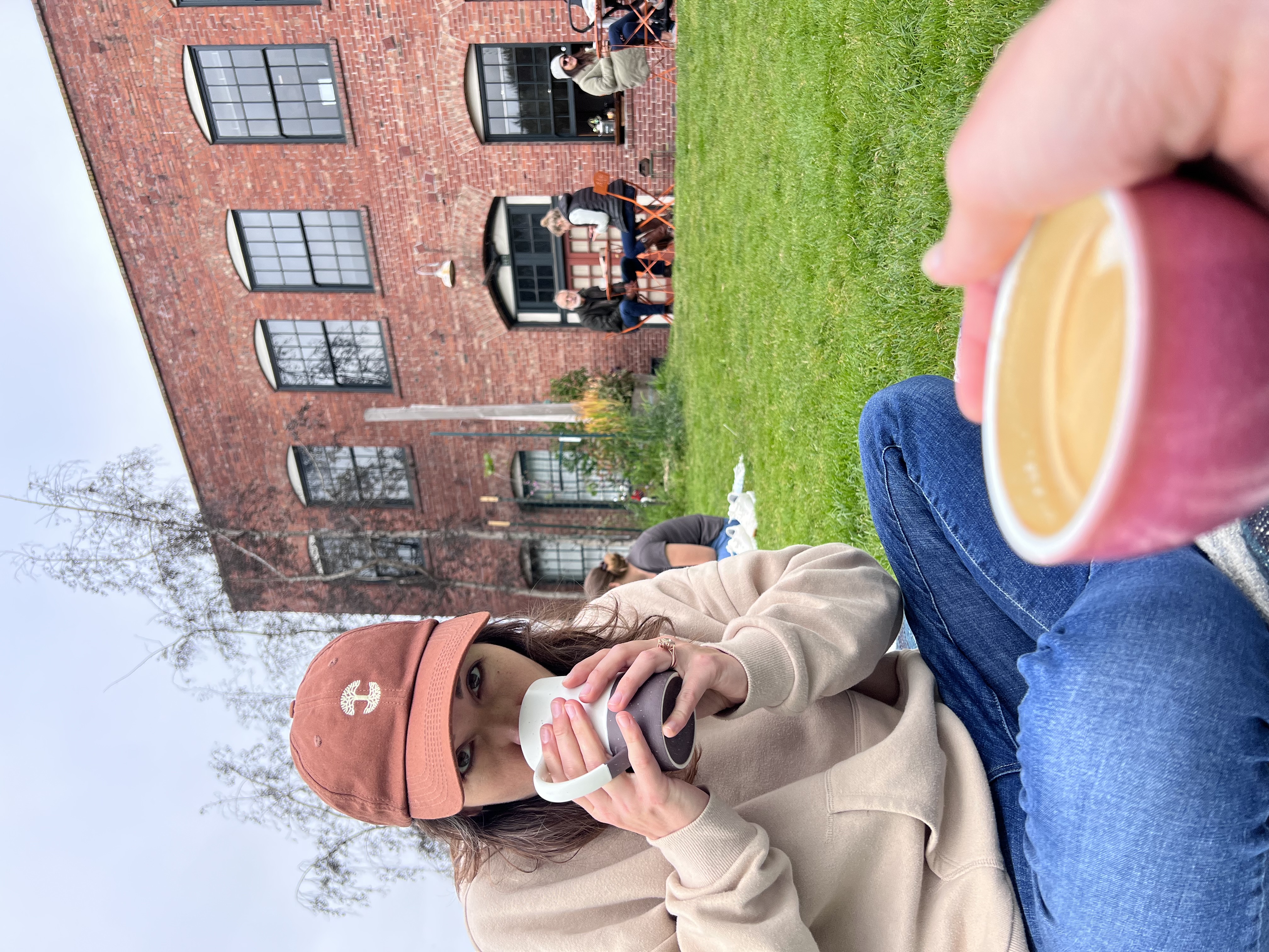 A woman in a rust-colored baseball cap and beige sweater sits on grass