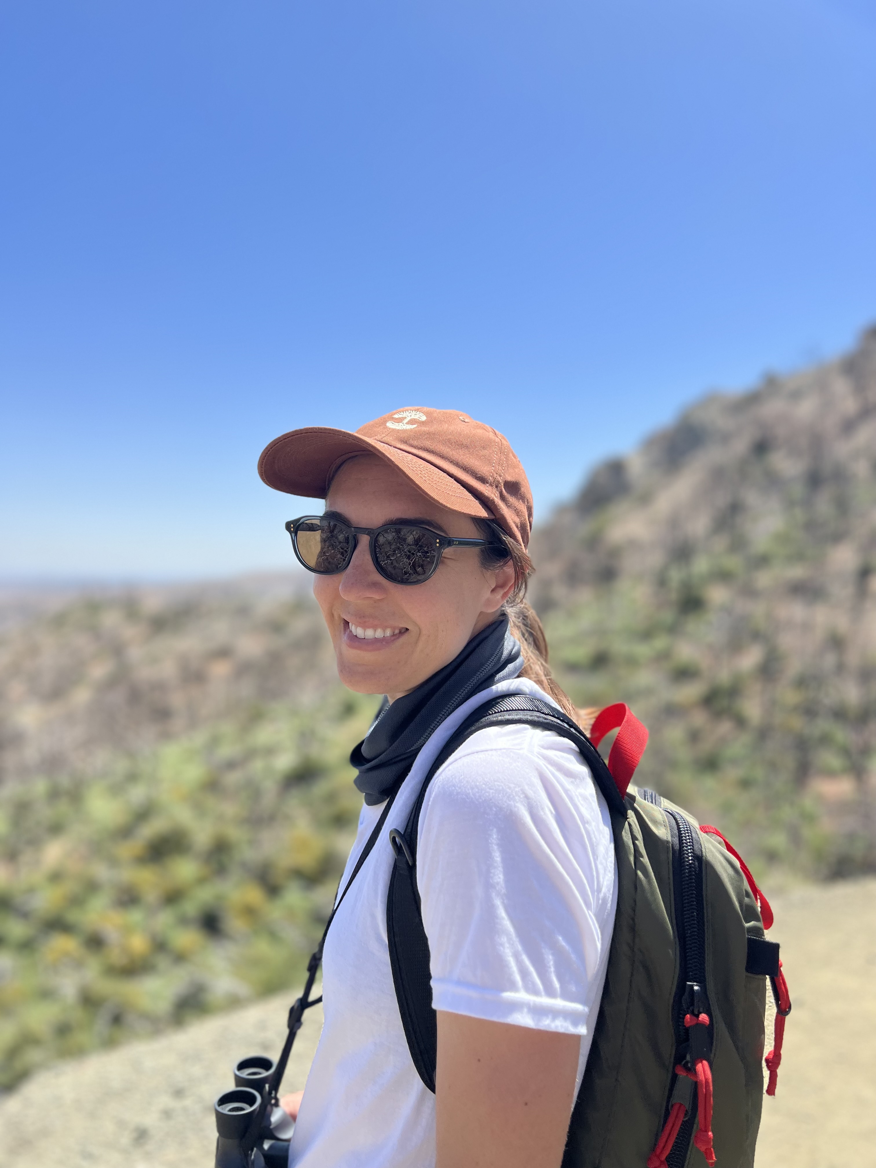 A smiling woman wearing sunglasses, an orange baseball cap, and a backpack