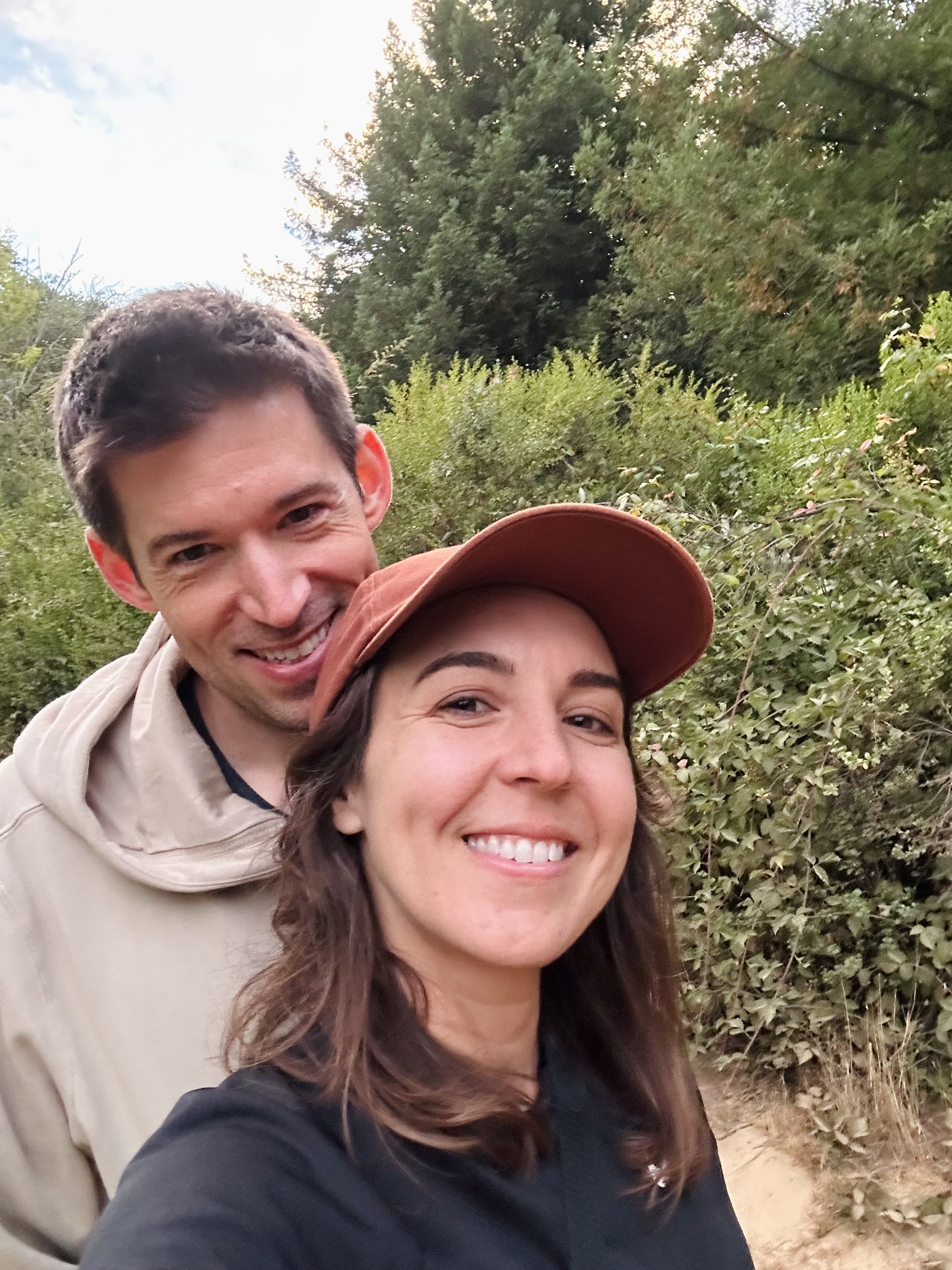 A smiling couple takes a selfie outdoors, with the woman wearing a brown
