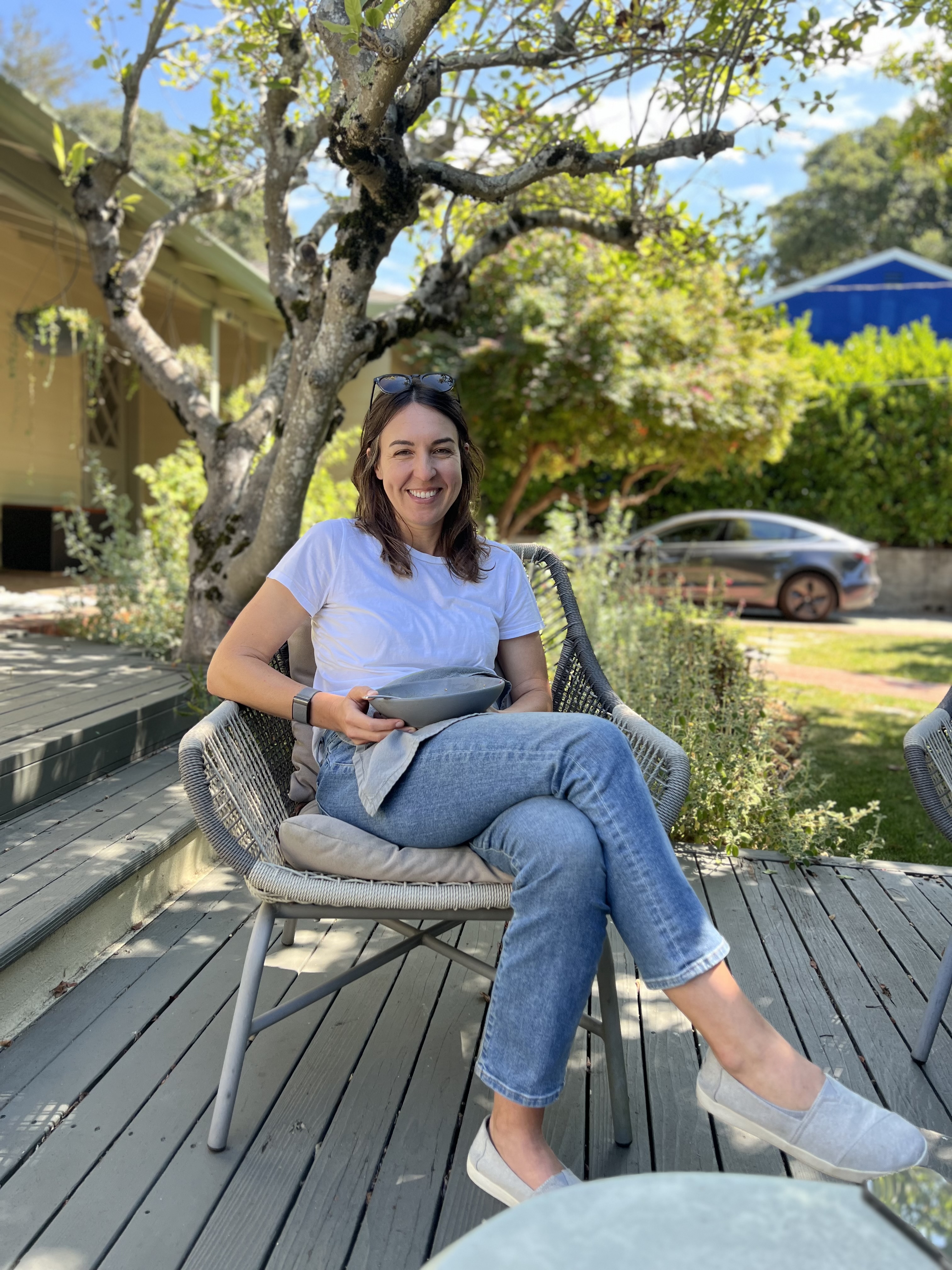A woman in a white t-shirt and jeans sits smiling in a wicker chair on a