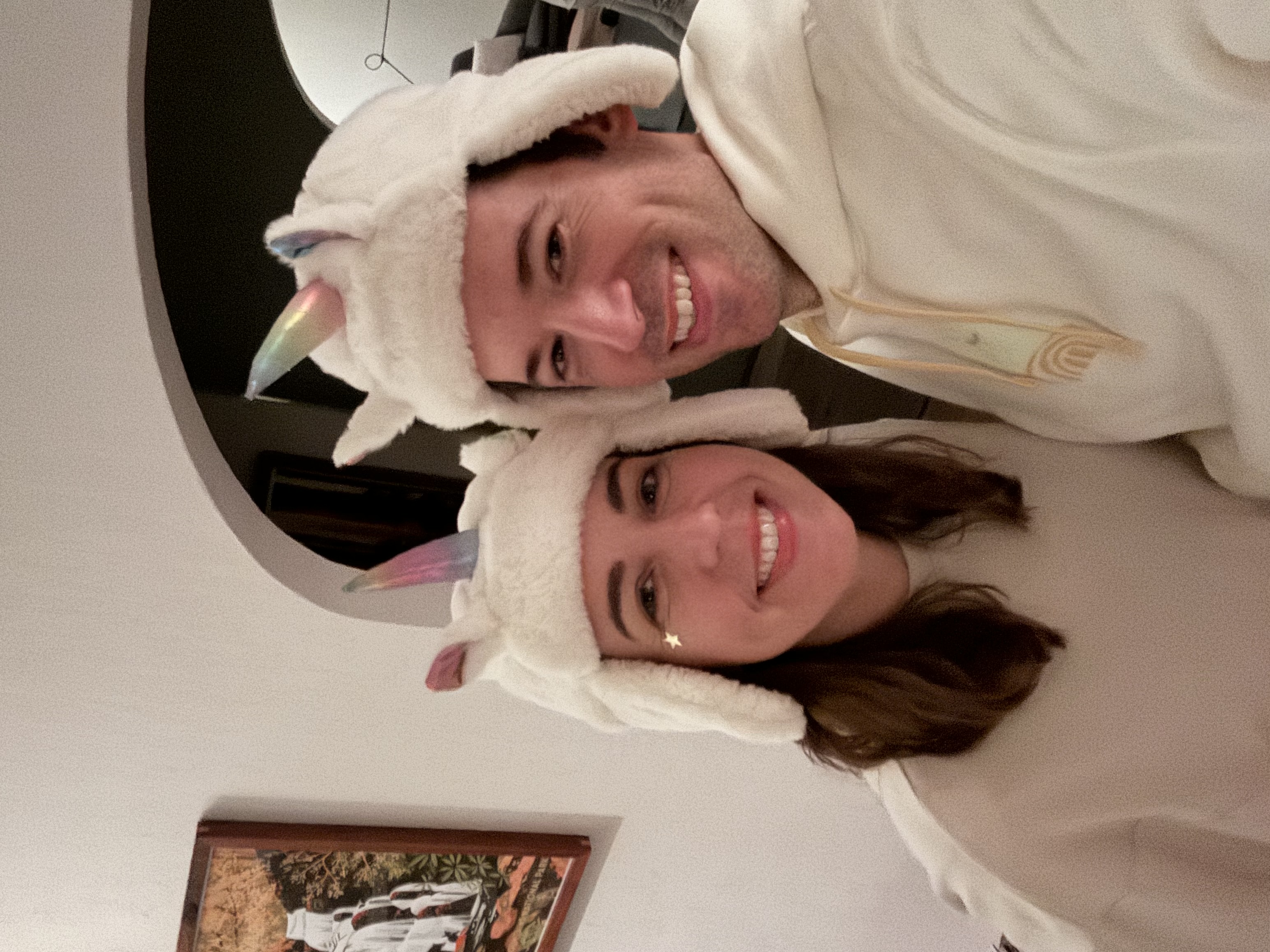 Two people wearing matching white unicorn hats with ears and horns smile at