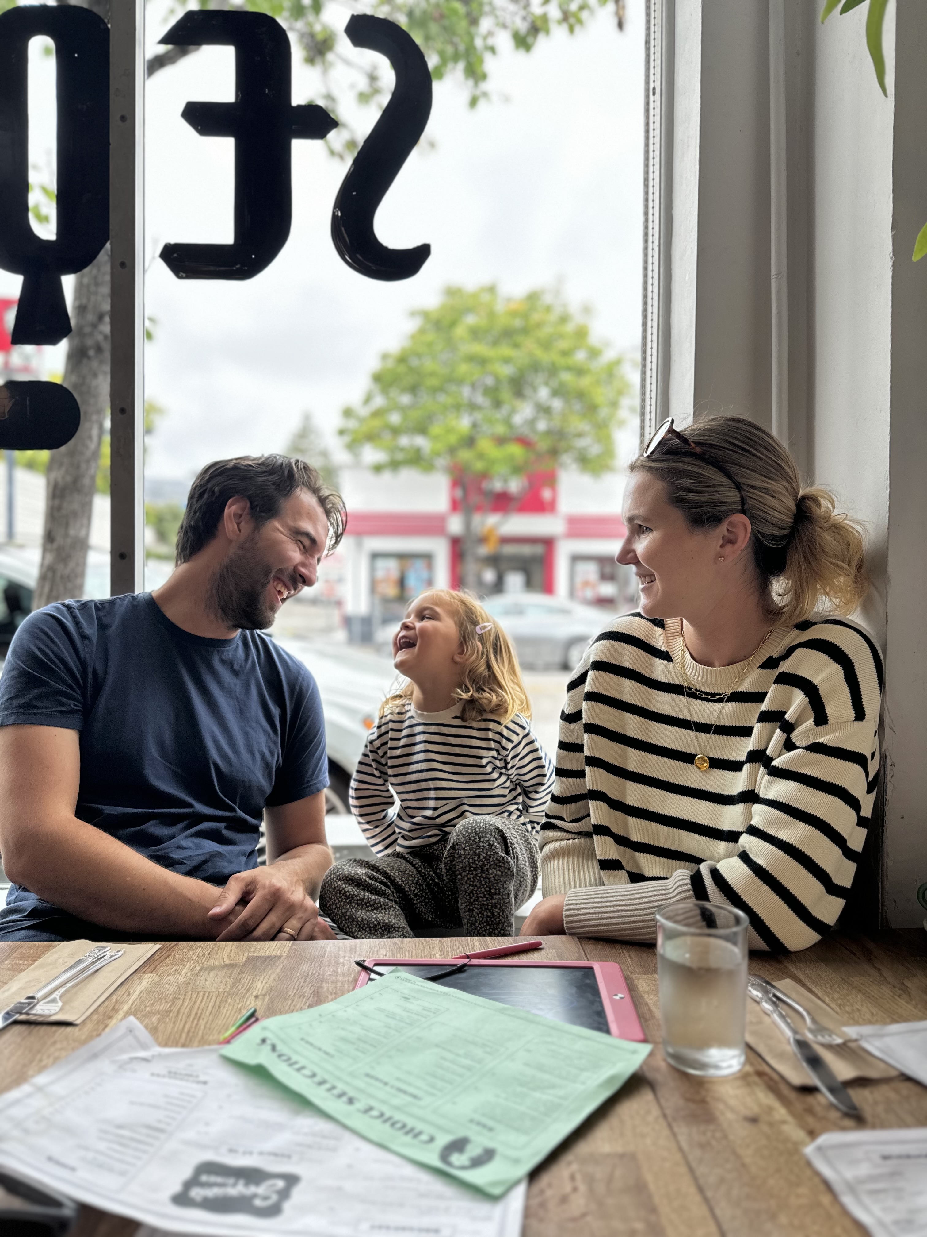 A happy family of three sits at a cafe table by a large window, with