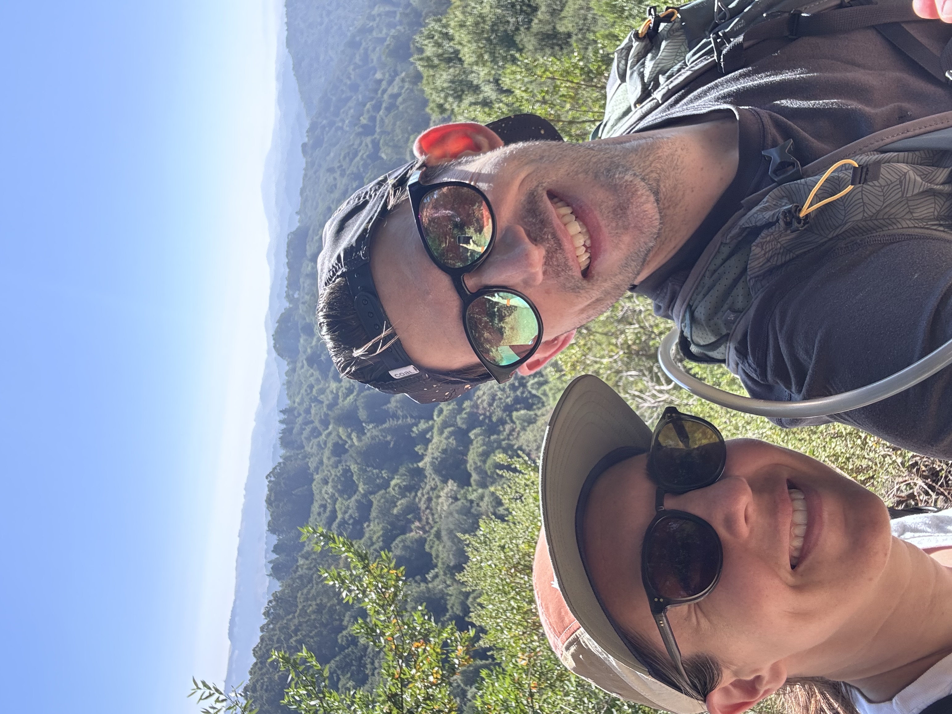 Two hikers wearing sunglasses and caps smile at the camera while taking a