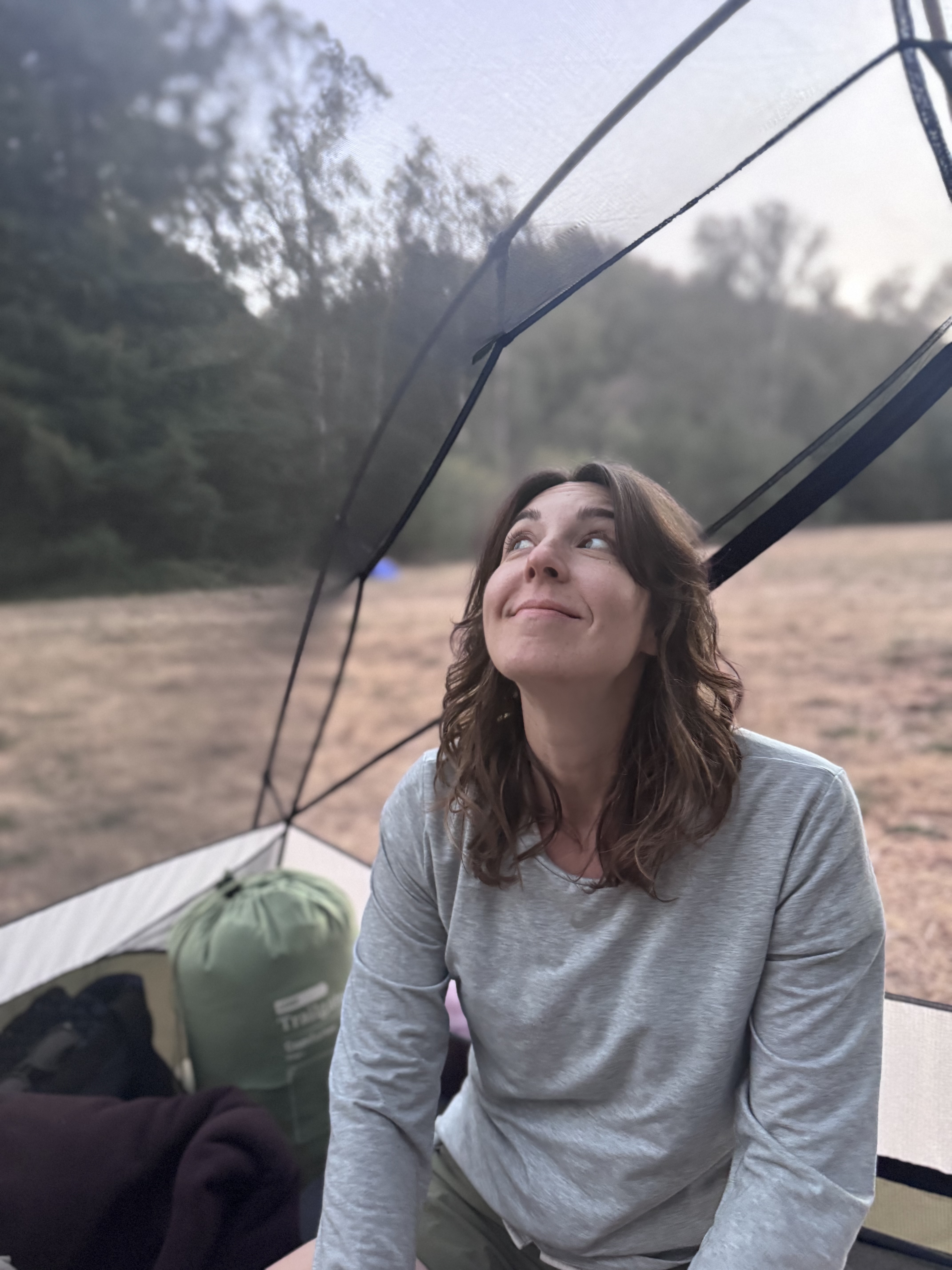A woman in a gray long-sleeved shirt sits inside a tent, looking up with a