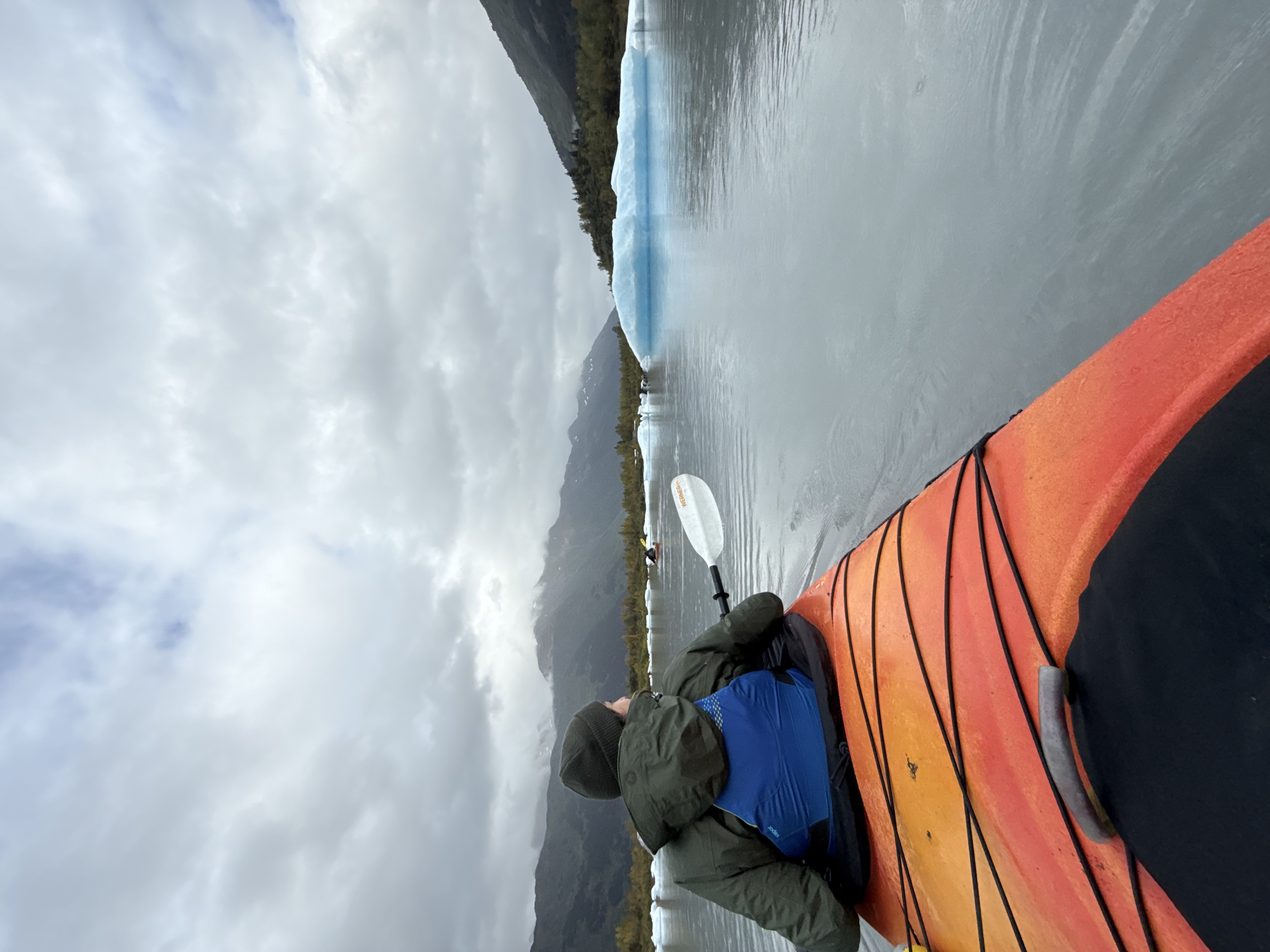 A kayaker in an orange kayak paddles through calm waters dotted with blue
