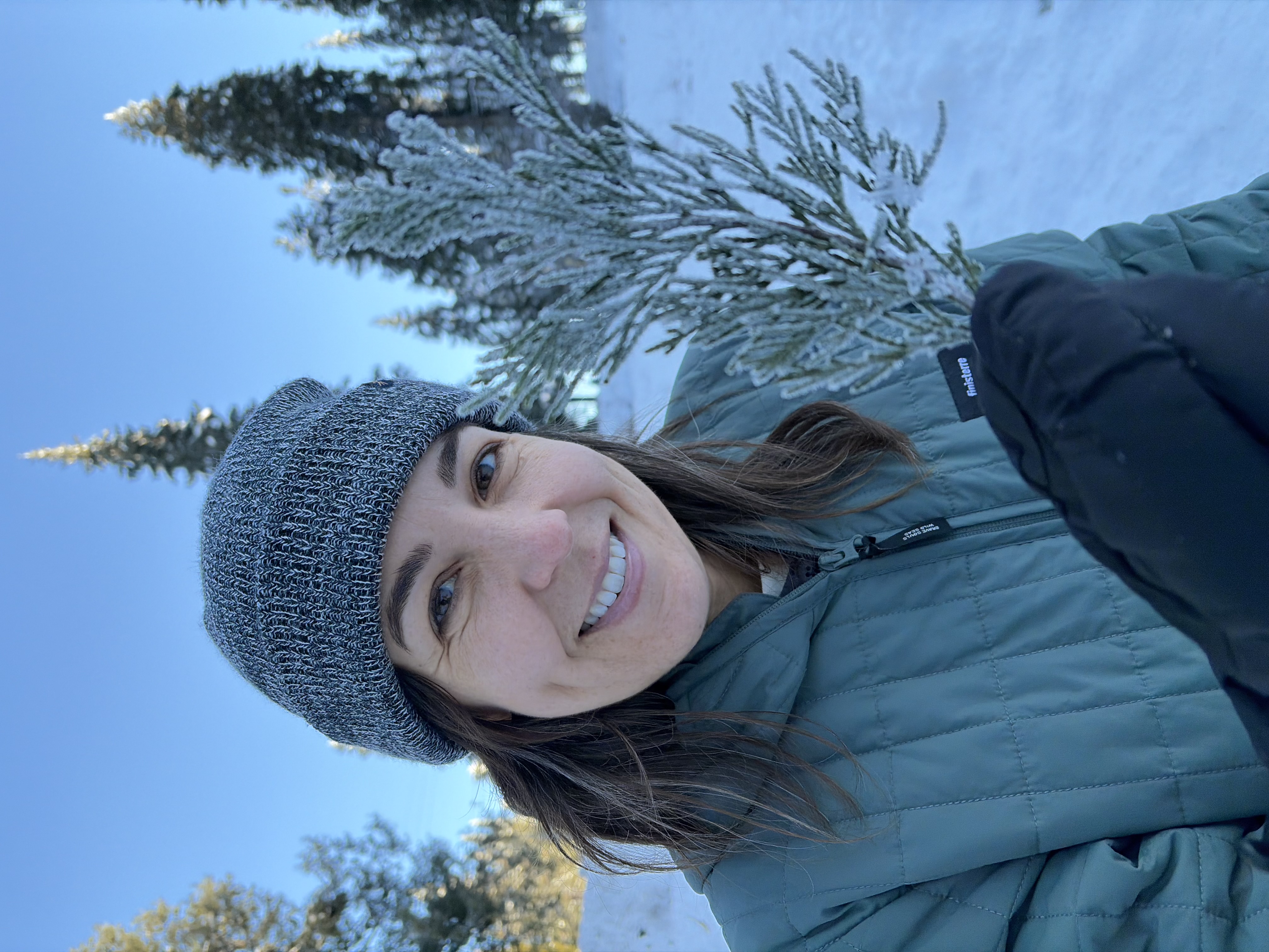 A smiling woman in winter clothing and a knit beanie holds a snow-covered